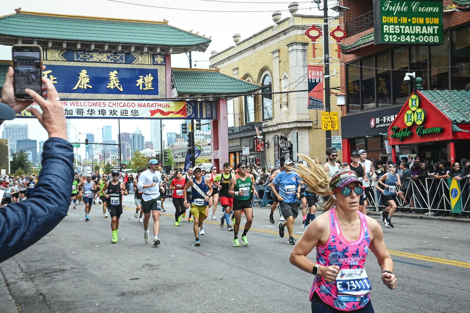 Runners passing under the Chinatown gate at Mile 21 of the Chicago Marathon with the downtown skyline visible behind and a spectator filming with their phone
