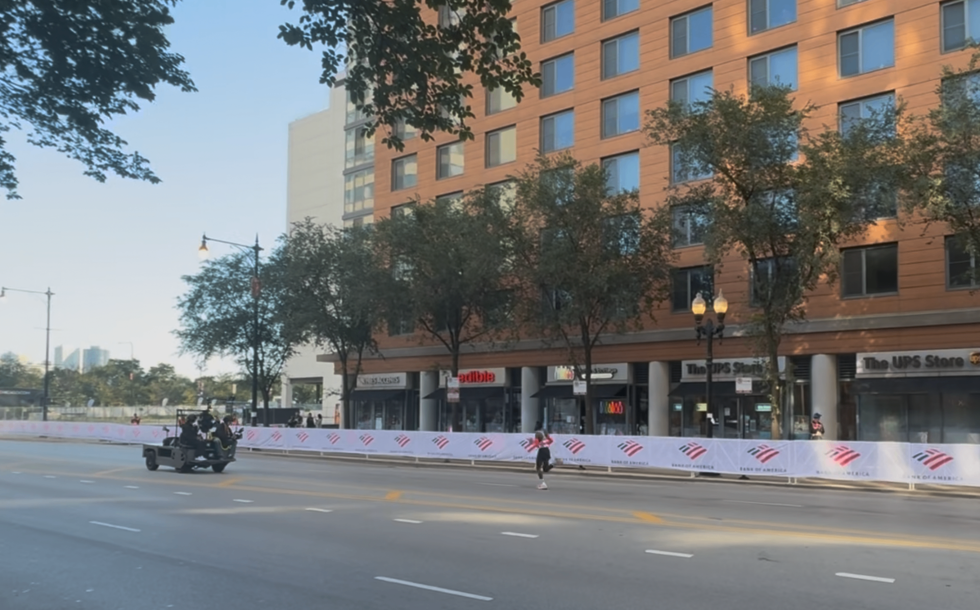An elite runner approaching the Mile 25/26 stretch of the Chicago Marathon trailed by a Bank of America camera cart with spectators cheering behind the barricades