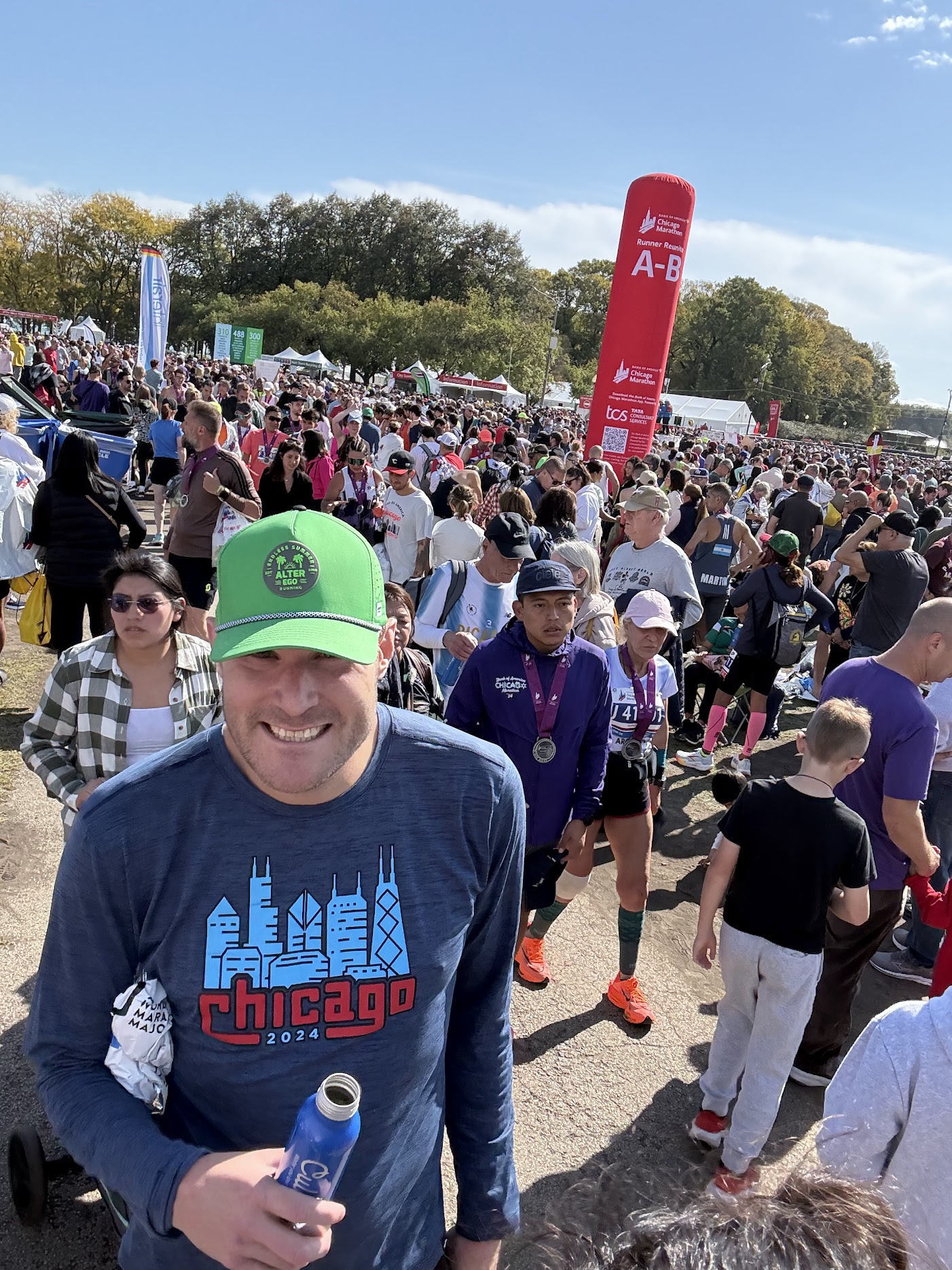 A finisher in a Chicago 2024 long-sleeve shirt walking through the crowded Runner Reunite area in Grant Park, with the red A-B letter tower visible behind the crowd